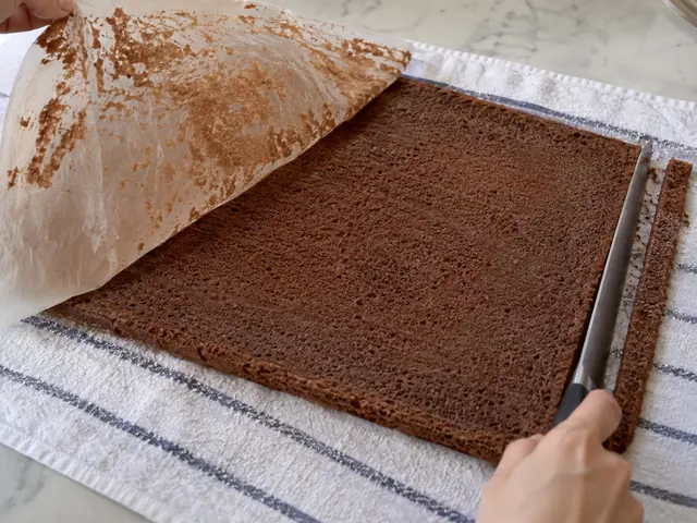 A thin chocolate sponge cake just turned out onto a kitchen towel dusted with sugar, the waxed paper being peeled away, and a knife trimming the edge