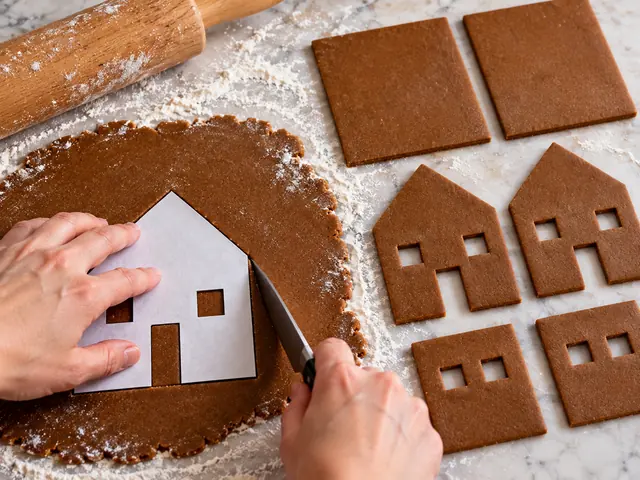 Rolled-out gingerbread dough on a floured counter with a paper pattern laid on top, a knife cutting out a house front with a pointed peak, cut wall and roof pieces nearby
