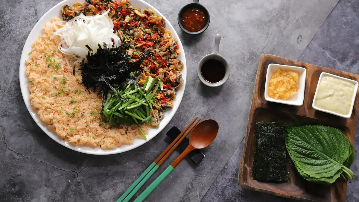Spicy clam bibimbap platter with rice, seasoned clams, garlic chives, onion and seaweed, with perilla leaves and dipping sauces